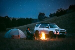 Man Sitting by Campfire and Silver Toyota with GOBI Racks Roof Rack on Top