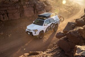 White off-road SUV with rooftop gear and a bike drives a dusty canyon trail at sunset.