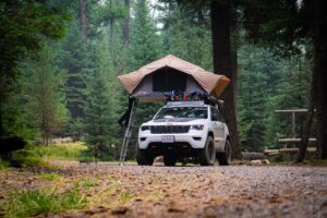 A white Jeep equipped with a rooftop tent is parked in a forest campsite